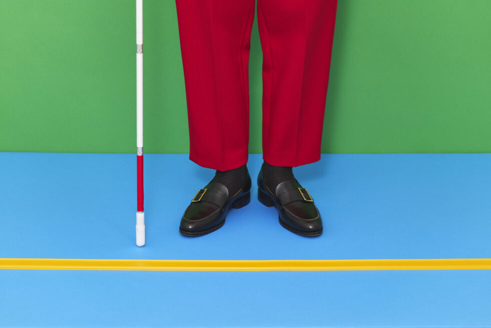 ココテープのビジュアル。背後の壁が緑、床は水色で黄色いココテープが横にまっすぐ引かれている。ココテープに両足を揃えて赤いズボン、黒いローファーを履き、白杖を持った人が立っている。