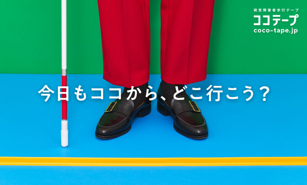 ココテープのビジュアル。背後の壁が緑、床は水色で黄色いココテープが横にまっすぐ引かれている。ココテープに両足を揃えて赤いズボン、黒いローファーを履き、白杖を持った人が立っている。