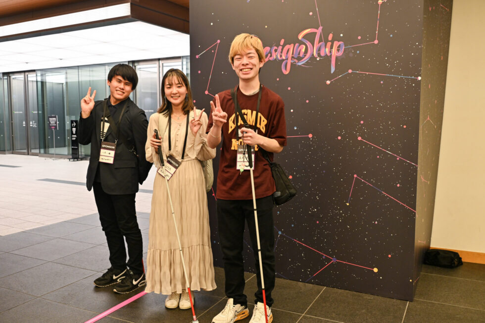 Designshipの看板の前で記念写真を撮る、視覚に障害のある学生３名。みんな笑顔でピースをしている。