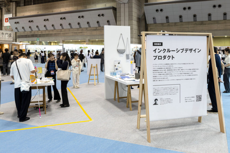 東京ビッグサイトの特設ブース「インクルーシブデザインプロダクト」に多くの来場者が集まっている。正面には大きな説明パネルが設置され、左手では来場者が視覚障害をテーマにしたプロダクトを手にして会話をしている。明るく開放的な空間に、多様な人々が集まっている様子が伝わる。