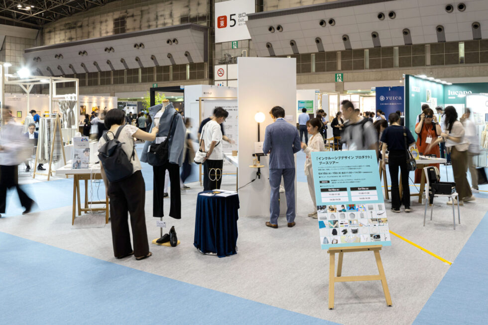 「インクルーシブデザインプロダクト」ブースの様子。ジャケットを着たマネキンや照明器具と一体となった手すりなどが展示されている。中央にはブースツアーのスケジュールが書かれた案内板があり、背後には多くの人が交流しながら製品を体験している様子が見える。