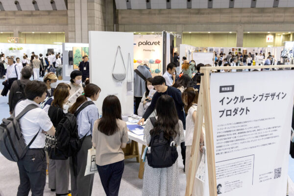 展示会場の「インクルーシブデザインプロダクト」ブースに、多くの来場者が集まり、PLAYWORKSタキザワの説明を受けている様子。壁面にはグレーのショルダーバッグが展示されている。会場全体が賑わいを見せている。