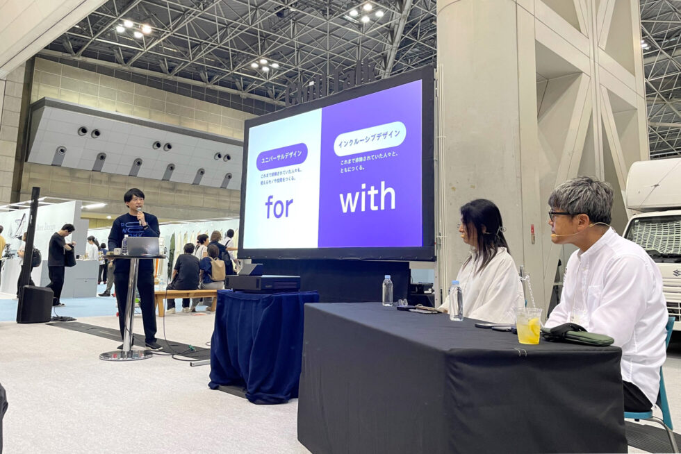 トークセッションの様子。PLAYWORKSタキザワがスクリーンに映ったスライドを使用して、インクルーシブデザインについて説明している。手前の机には、フェリシモ 筧さん、コクヨ 藤木さんが座っている。
