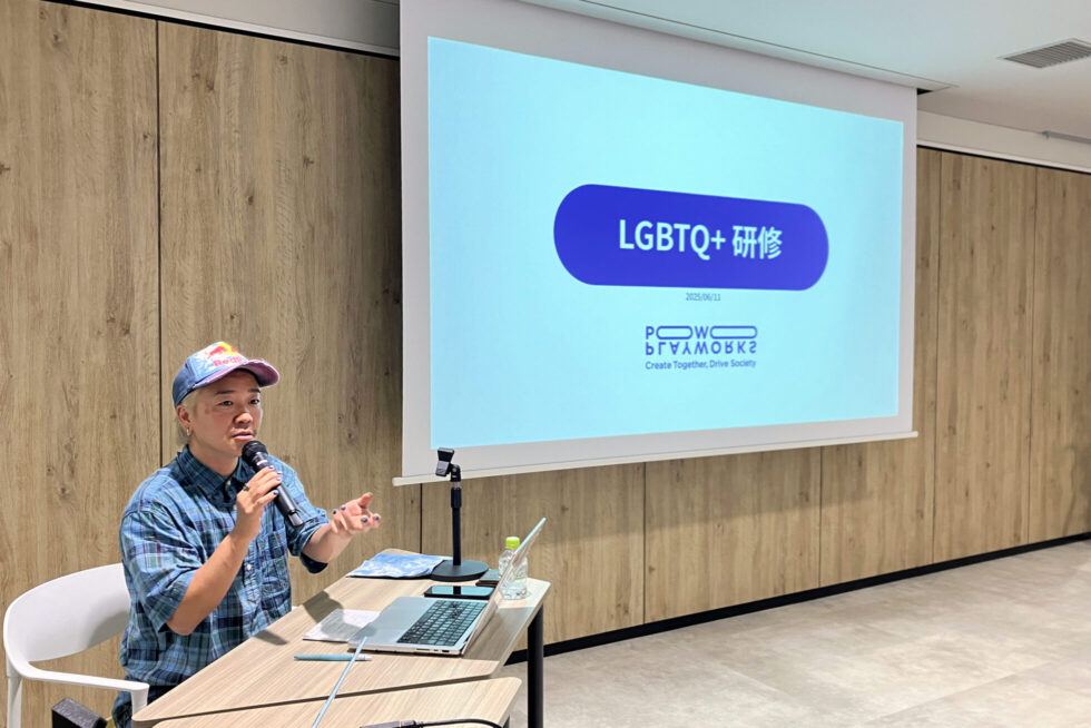 マイクを手に持ち話をする講師の大嶋さん。チェック柄のシャツとカラフルなキャップを着用し、背景のスクリーンには「LGBTQ+研修」と日付（2025年6月11日）が表示されている。