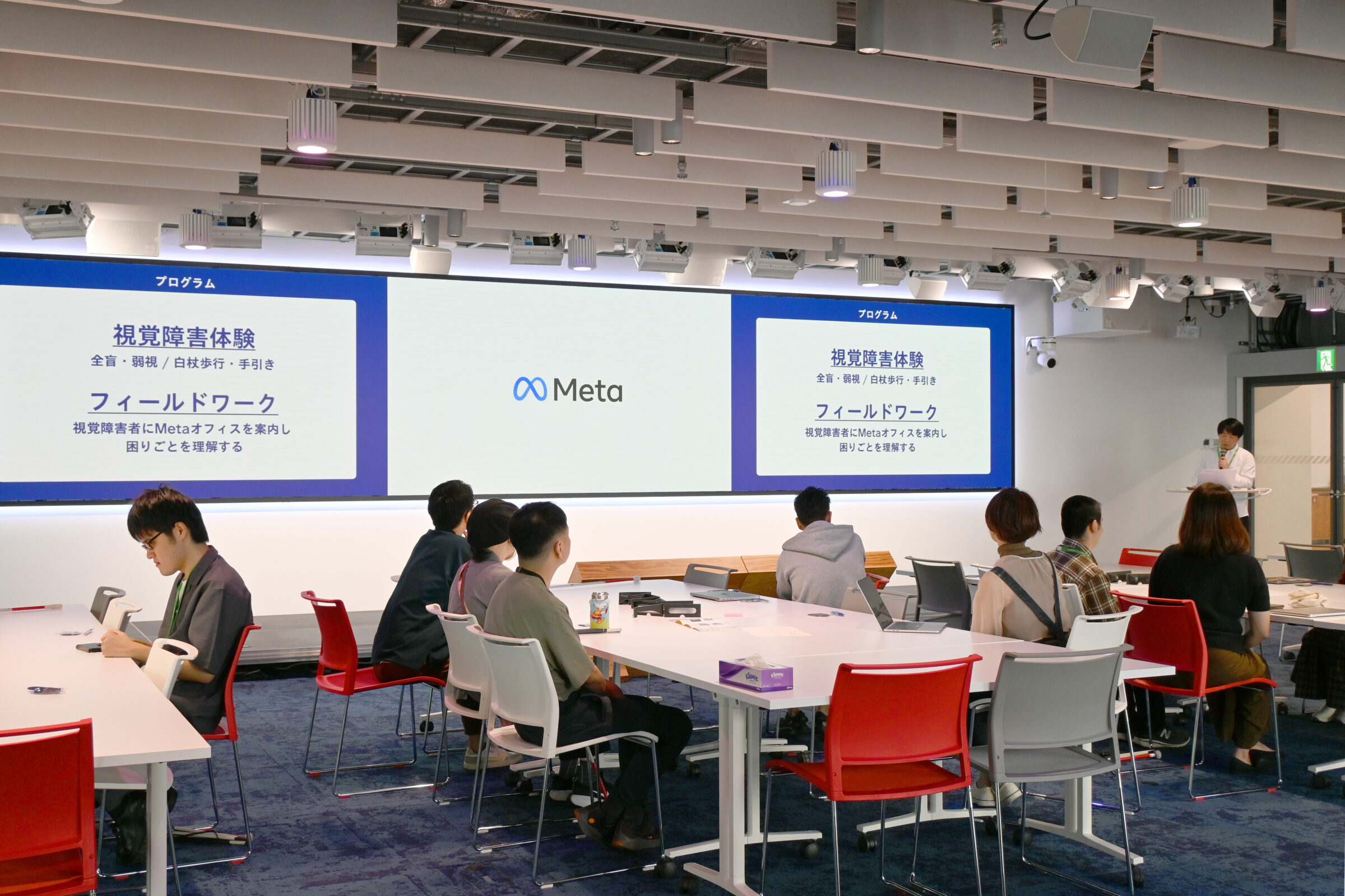 大型スクリーンに「視覚障害体験」「フィールドワーク」とプログラムが映し出され、参加者が説明を聞いている。