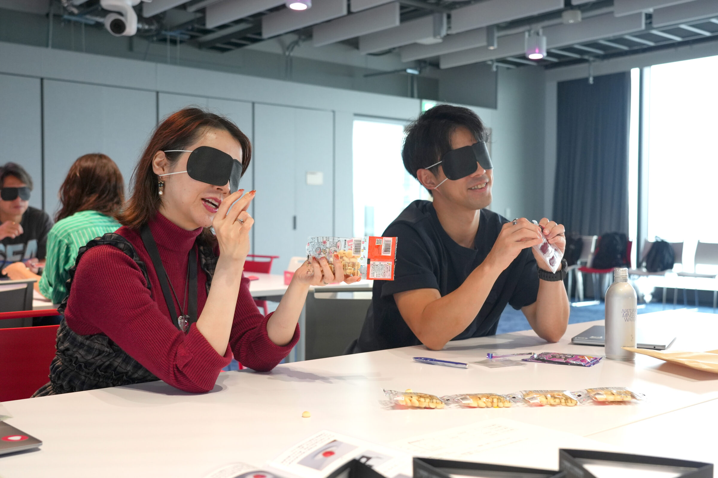 アイマスクを着けた2人の参加者が、お菓子の袋や中身を手に取り、触覚などを頼りに種類を確かめている様子。笑顔で会話しながら体験している。