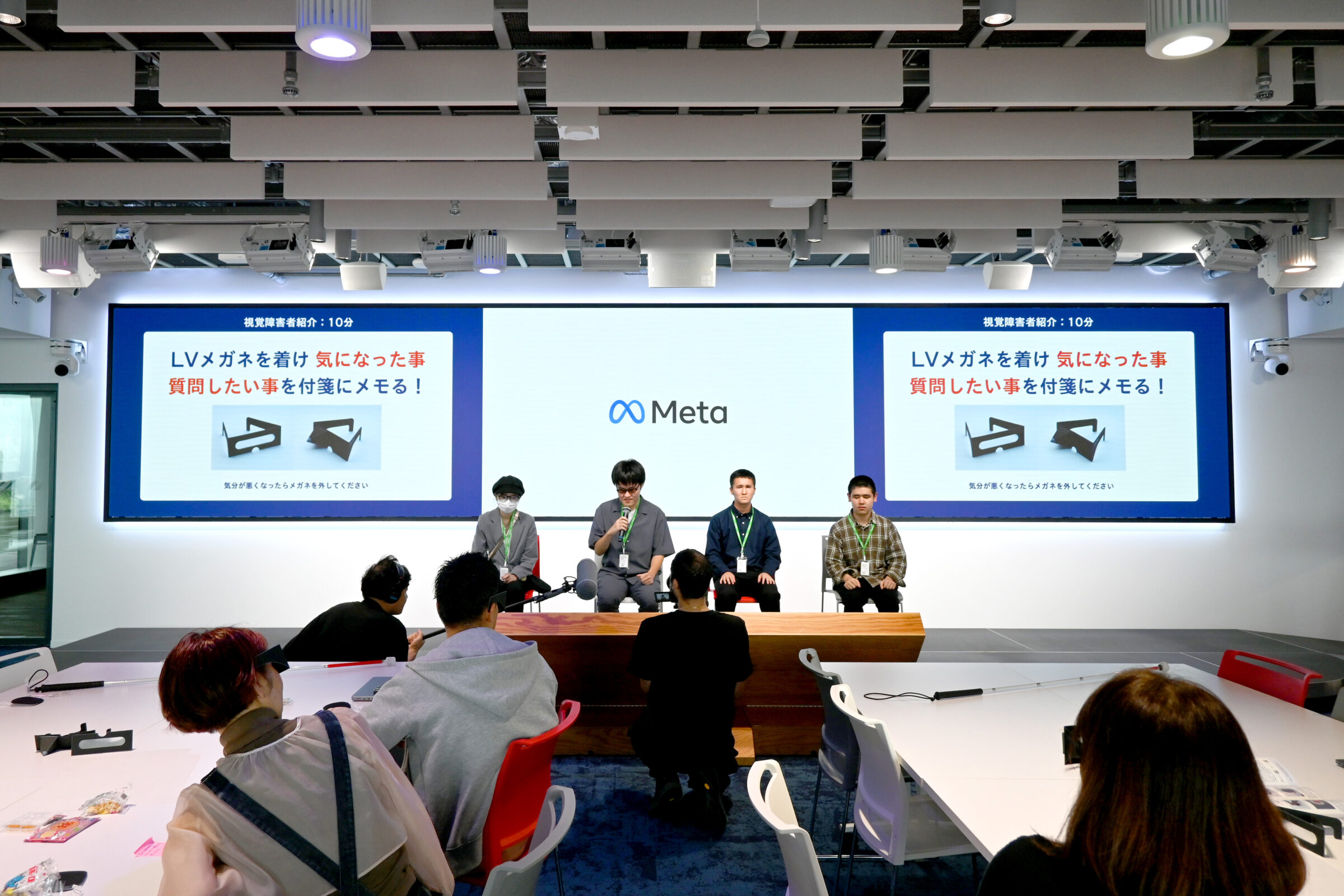 会場前方に4人の視覚障害リードユーザーが並び、参加者がロービジョン体験眼鏡を着けながら話を聞いている。スクリーンには「ロービジョンメガネを着け 気になった事・質問したい事を付箋にメモる！」と表示されている。