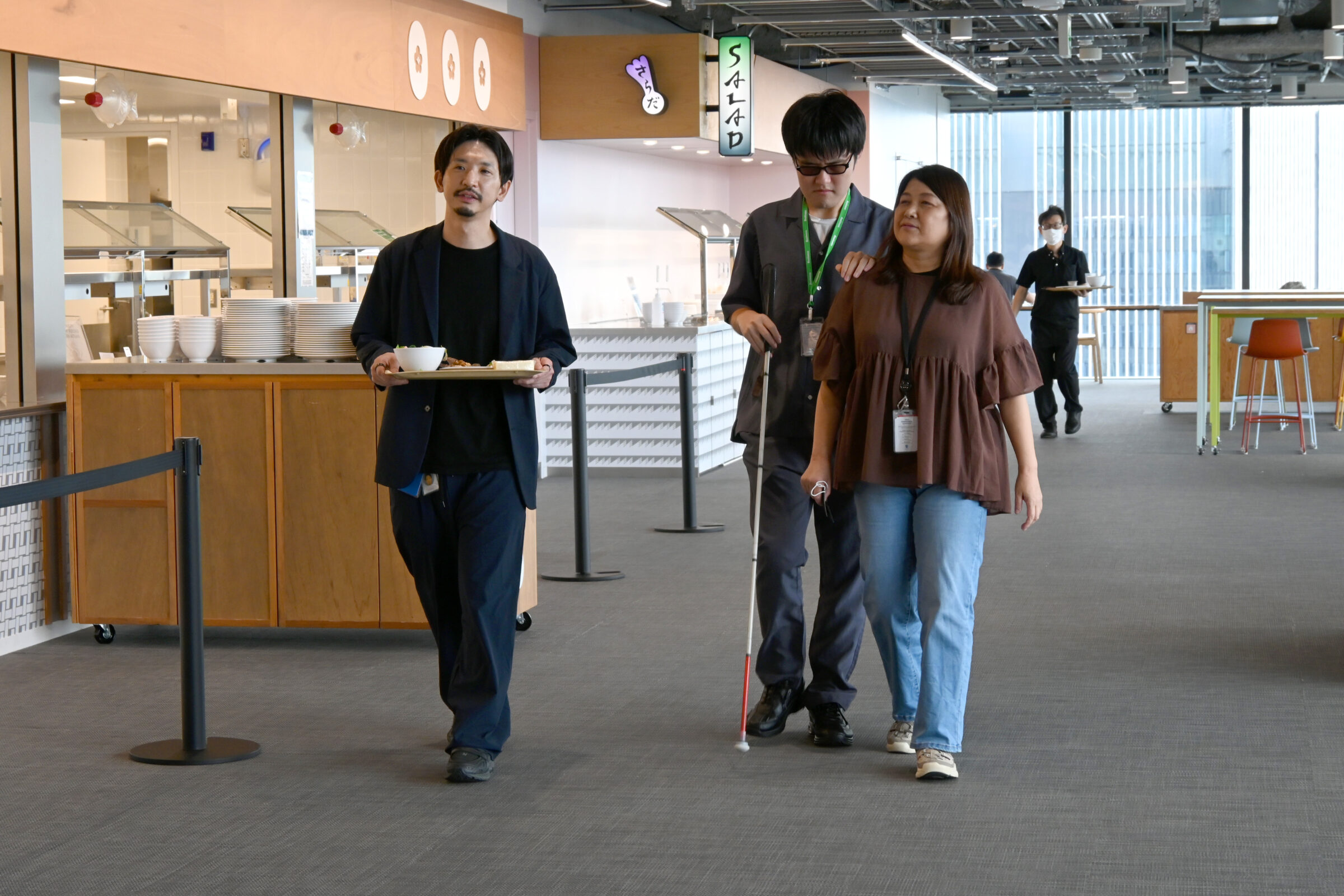 Metaオフィスの食堂で、白杖を持つ視覚障害のあるリードユーザーが同行者の肩につかまり、料理を載せたトレーを持つ参加者と一緒に移動している様子。