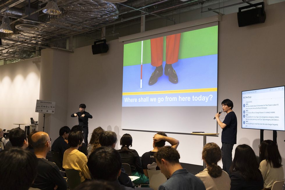 PLAYWORKS代表のタキザワケイタが、視覚障害者の移動に関するスライドを投影し、手話通訳者とともにプレゼンテーションを行っている。